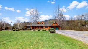 A two-story log cabin with a green garage, surrounded by a well-maintained lawn and trees. - Sycamore Cabin with Hot Tub Breathtaking Views (Gaines)