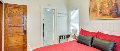 A bedroom with a red bedspread, a wooden door, a ceiling fan, and a painting on the wall.