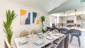 A modern dining area with a long table set for six, surrounded by chairs. There are two large abstract paintings on the wall, a potted plant, and a kitchen area visible in the background. - Game Room, Hot Tub & Mtn Views: Palm Springs Gem (Palm Springs)