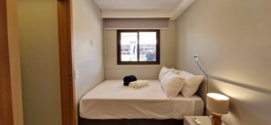 A small, neatly arranged bedroom with a single bed, a nightstand, a lamp, and a window with a view of a bridge. - COARM0104 - Smart Convenções (Salvador)
