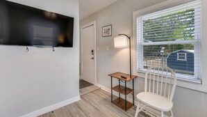 Una habitación con un televisor de pantalla plana montado en la pared, una mesita con una lámpara, una silla blanca y una ventana con persianas.