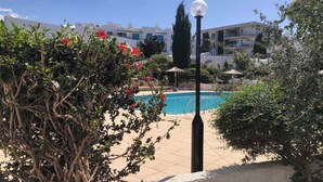 A pool area with a clear blue sky, surrounded by greenery and white buildings with balconies. - Venus Beach | Elegant 1BR Apt | 100m From Sea | Groundfloor (Paphos)