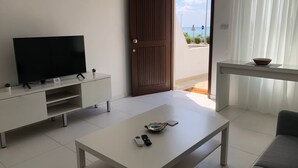 A modern living room with a flat-screen TV, a coffee table, and a sofa. A wooden door leads to a balcony with a view of the sea. - Venus Beach | Elegant 1BR Apt | 100m From Sea | Groundfloor (Paphos)