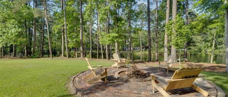 A circular fire pit with wooden chairs and a bench, surrounded by a brick patio, set in a grassy area with tall trees and a body of water in the background.