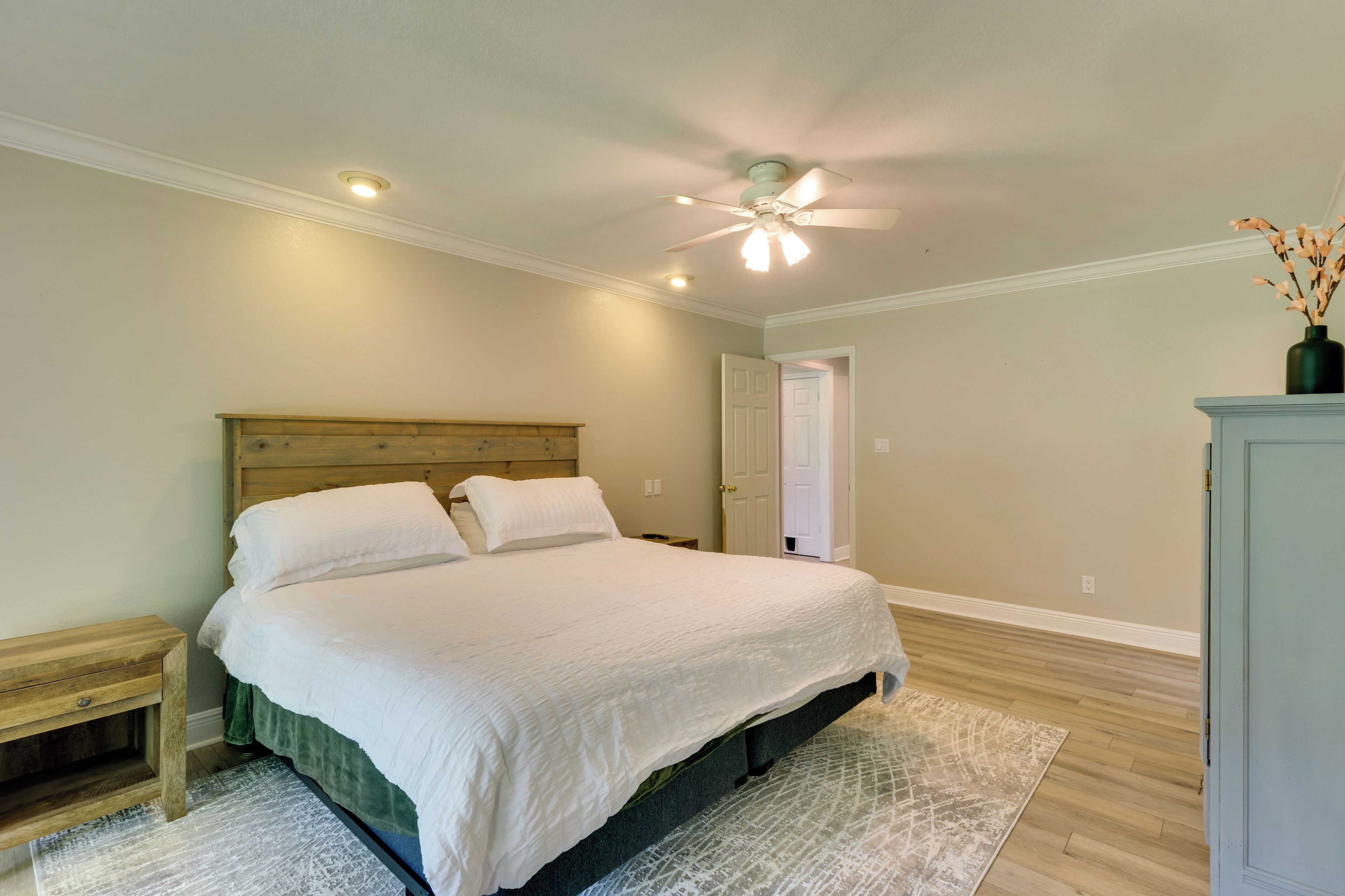 A bedroom with a bed, wooden headboard, bedside tables, a ceiling fan, and a door leading to another room.