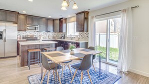 Egy modern konyha, központi szigettel, sötét színű, fából készült szekrényekkel, rozsdamentes acélból készült háztartási gépekkel, valamint egy kék mintás szőnyeggel díszített étkezővel.