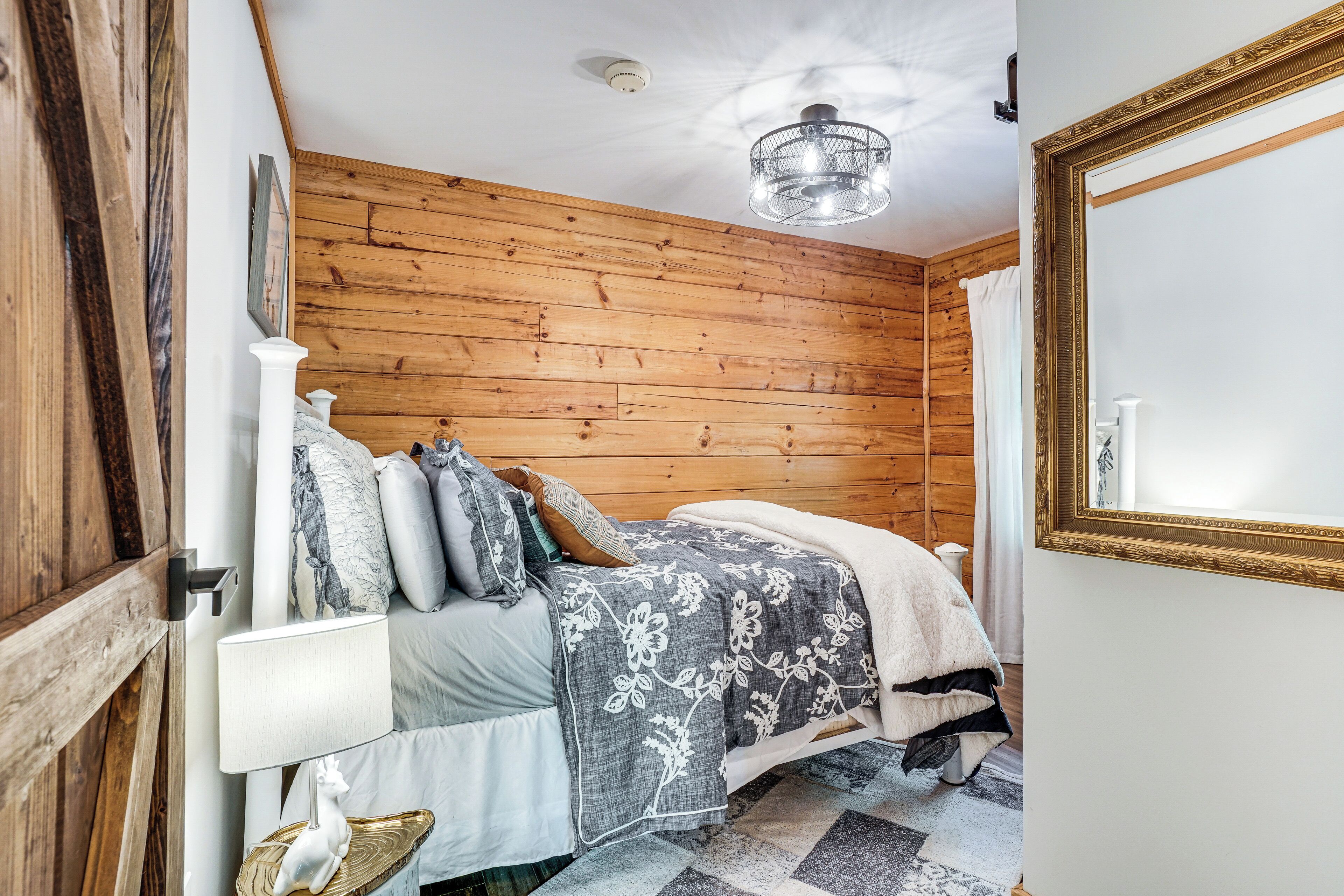 Un dormitorio con pared de madera, una cama con colcha floral, una lámpara de noche y un espejo con marco dorado.
