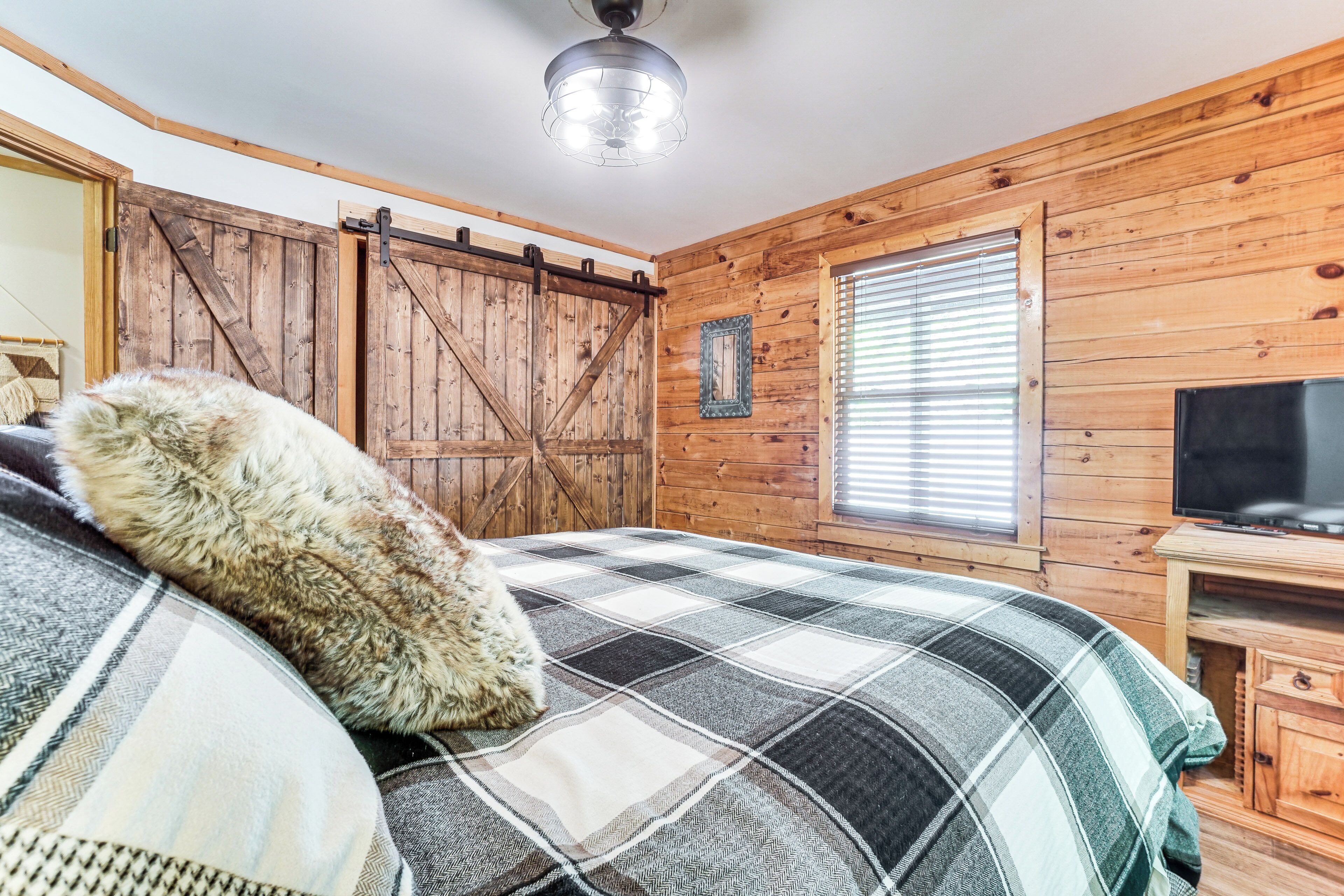 A cozy room with a plaid bedspread, wooden walls, a barn-style door, and a television on a wooden stand.