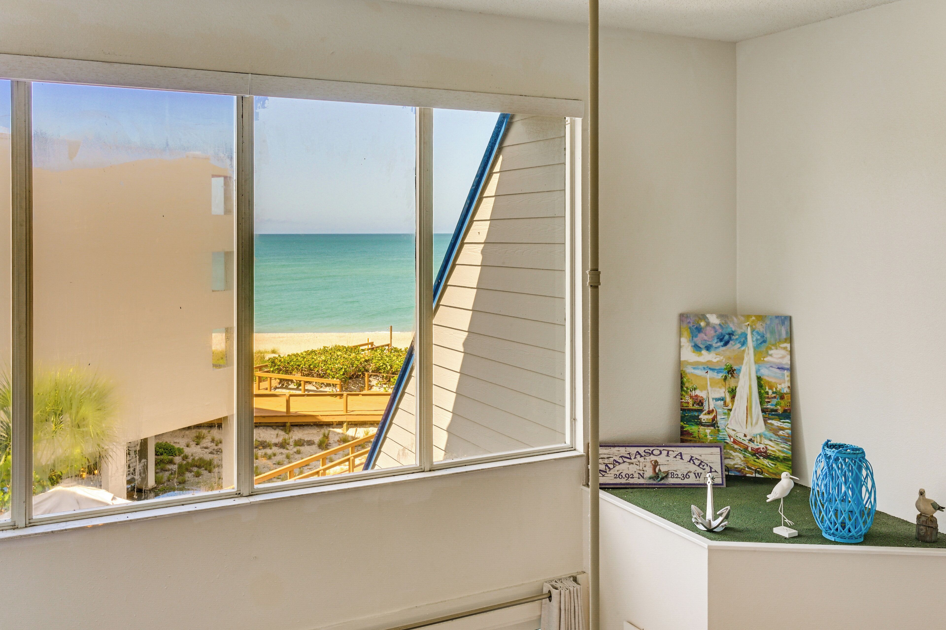 Una habitación con un ventanal grande con vistas a la playa, una mesita con objetos decorativos y un cuadro en la pared.