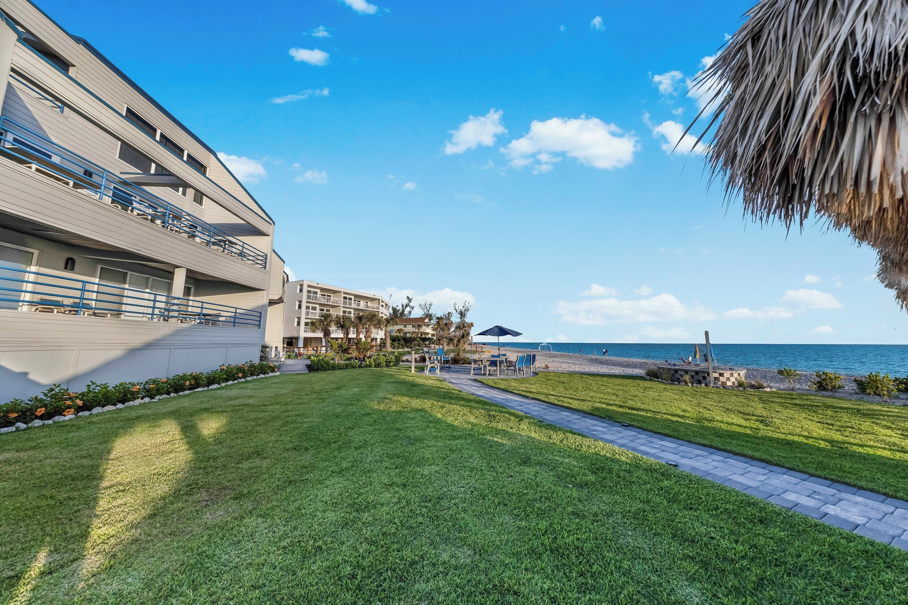 Un patio de hotel con un camino que lleva a una playa, una zona de piscina y un edificio con balcones.