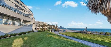 Un patio de hotel con un camino que lleva a una playa, una zona de piscina y un edificio con balcones.