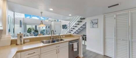 A modern kitchen with white cabinets, a stainless steel sink, and a dishwasher. There is a staircase leading to another level, and a view of the outdoors through large windows.