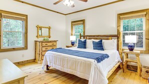 Ein Schlafzimmer mit einem großen Bett, einem Holzkopfstück, zwei Nachttischen mit Lampen, einer Kommode und einem Fenster mit Blick auf Bäume.