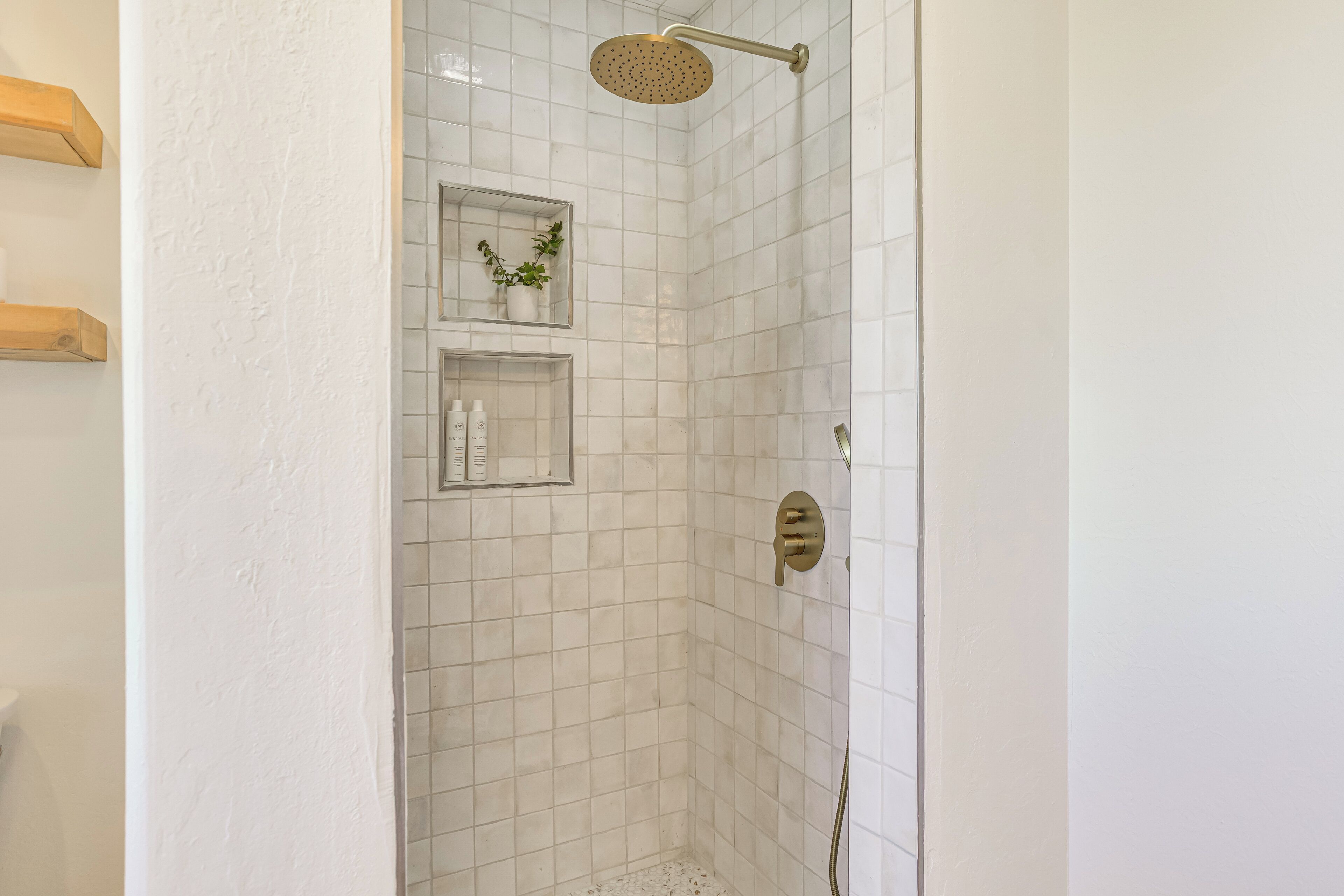 Une douche aux carreaux blancs, un pommeau de douche doré et une petite étagère avec des articles de toilette.