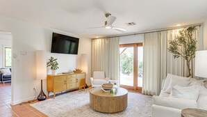 A modern living room with a round coffee table, a white sofa, a flat-screen TV mounted on the wall, and a ceiling fan. - Lake Lawtonka View: Mountaintop Medicine Park Home (Medicine Park)