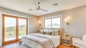 A bedroom with a large bed, a ceiling fan, a nightstand with a lamp, and a view through the window. - Lake Lawtonka View: Mountaintop Medicine Park Home (Medicine Park)
