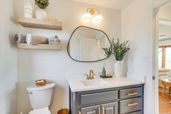 A bathroom with a white toilet, a white sink with a gold faucet, a mirror, and a shelf with towels and a basket.