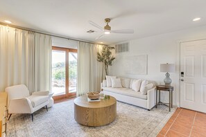 Una sala de estar con una mesa redonda de madera, un sofá blanco, un sillón blanco, una mesita de noche con una lámpara y un ventilador de techo.