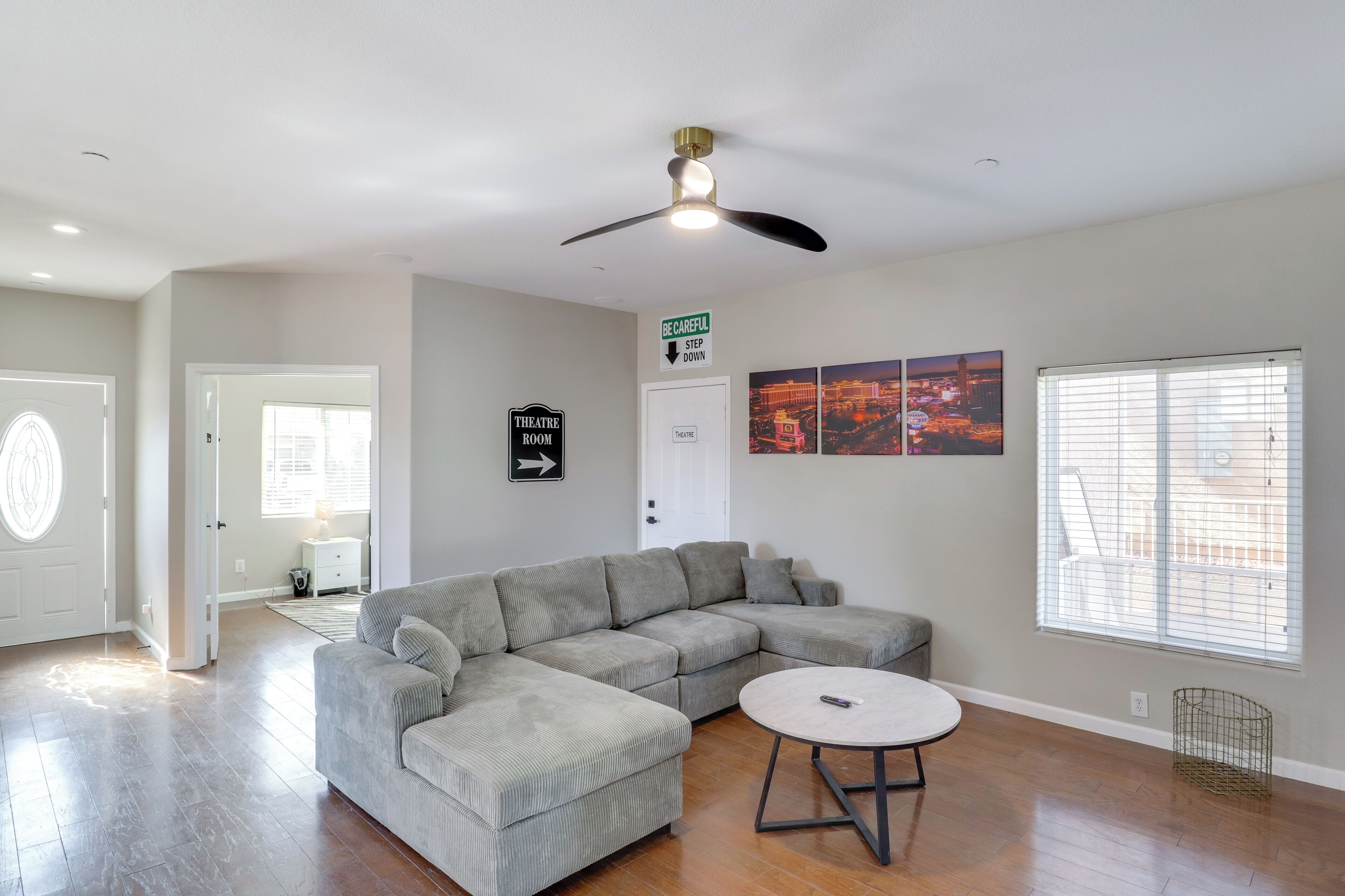 A spacious living room with a sectional sofa, a round coffee table, and a ceiling fan. A door with a sign points to the theatre room.