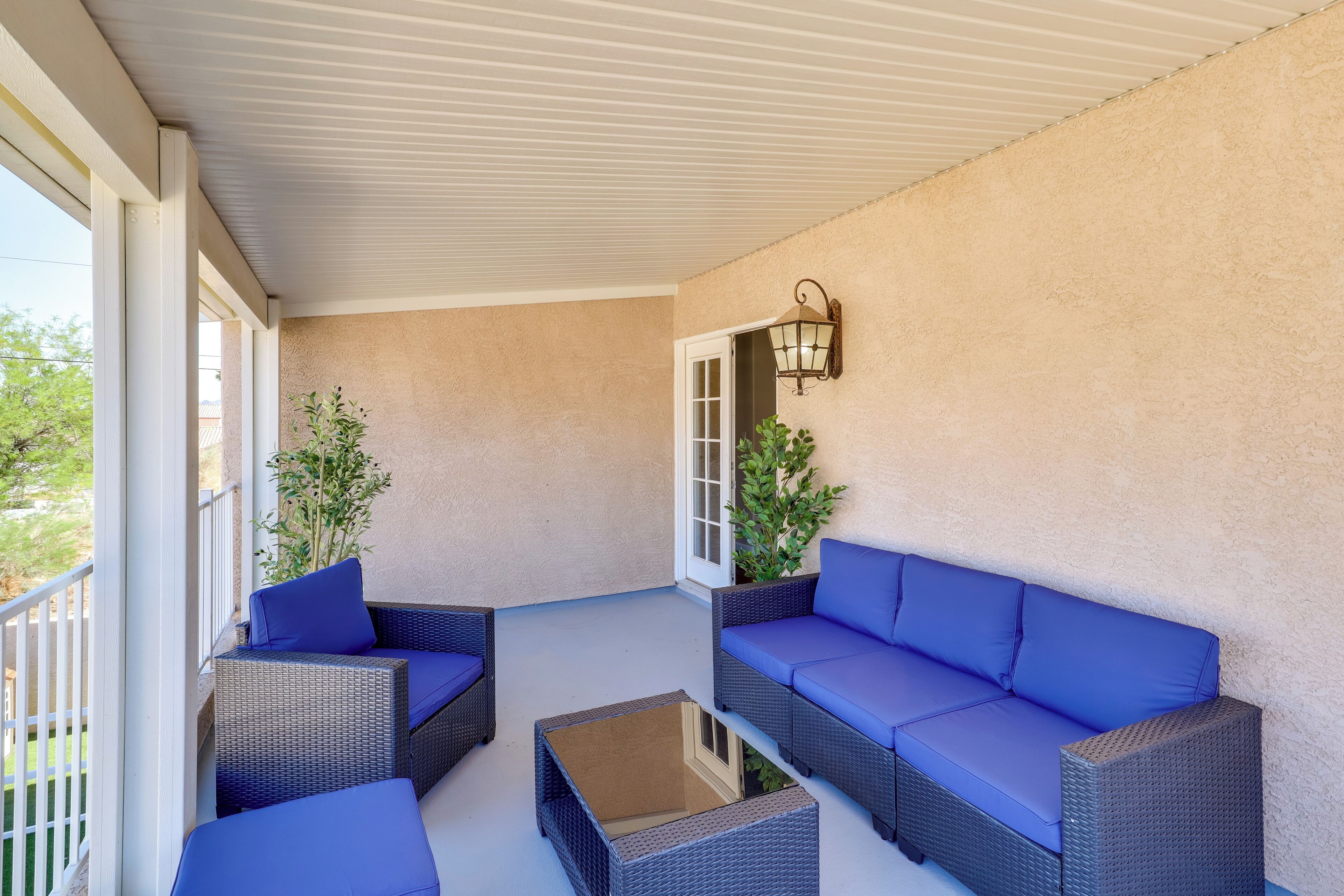 Un patio cubierto con mobiliario de mimbre y cojines azules, una mesa de centro de vidrio y un farol colgante en la pared.