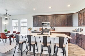 A modern kitchen with dark wooden cabinets, a large island with bar stools, a microwave, and a dining area with red chairs. - 6 Mi to Great Salt Lake: Mtn-view Retreat! (Tooele)