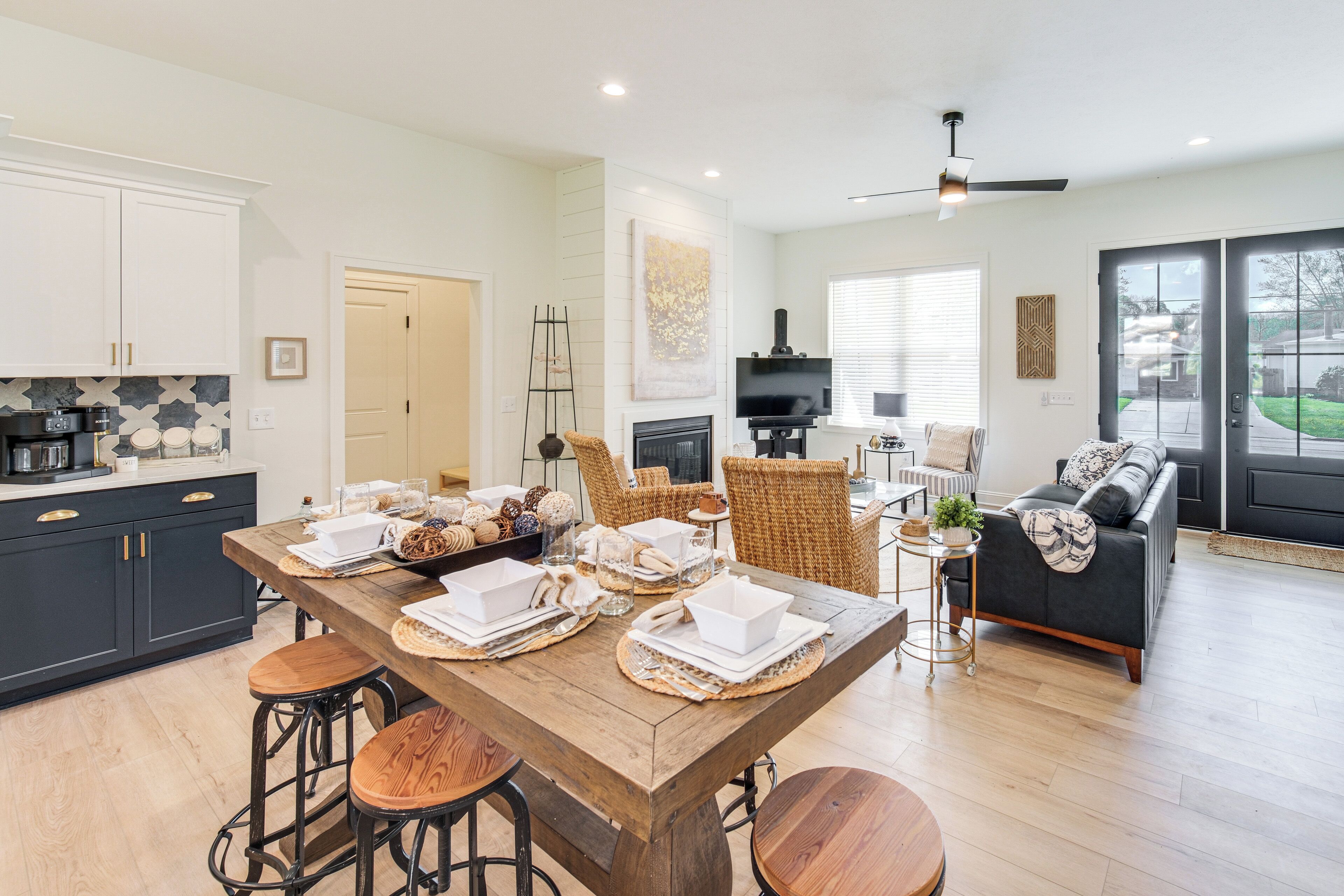 A modern kitchen with dark cabinets, a wooden island, and bar stools. There is a dining area with a sofa and chairs, and a fireplace.