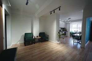 Un salón espacioso con piso de madera, dos sillones verdes, una mesita redonda y una planta en maceta. Se ve un área de comedor con una mesa y sillas al fondo.