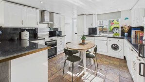 Une cuisine moderne avec des meubles blancs, un îlot central doté de chaises, un four encastré, un four à micro-ondes et un lave-linge.