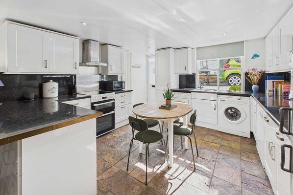 A modern kitchen with white cabinets, a central island with chairs, a built-in oven, a microwave, and a washing machine. - Host Stay Luna Cottage (Saltburn-by-the-Sea)