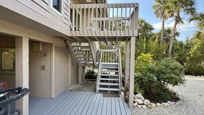 A two-story building with a wooden deck, a staircase, and a small garden area with palm trees and shrubs. - Black Skimmer Home | Relaxing Sanibel Escape, Walk to Beach (Sanibel)