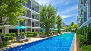 A swimming pool with lounge chairs and umbrellas, surrounded by greenery and multi-story buildings. - Title Rawai Tropical View Apartment B209 (Rawai)