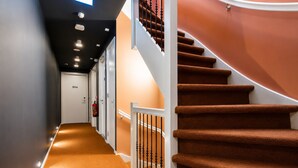 A hallway with a carpeted floor, a staircase with brown carpeted steps, and a fire extinguisher on the wall.