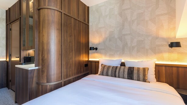 A modern hotel room with a large bed, a wooden wardrobe, a built-in mirror, and a patterned wall.