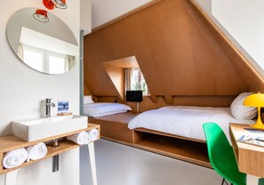 Ein modernes Hotelzimmer mit schrägem Dach, einem runden Spiegel, einem Nachttisch aus Holz mit Lampe, einem grünen Sessel und einem Schreibtisch mit Stuhl.