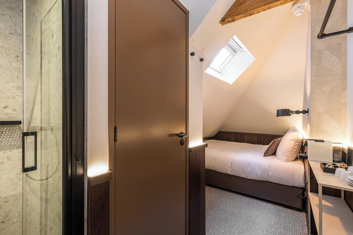 A small, cozy bedroom with a single bed, a bedside table with a lamp, and a window with a skylight.
