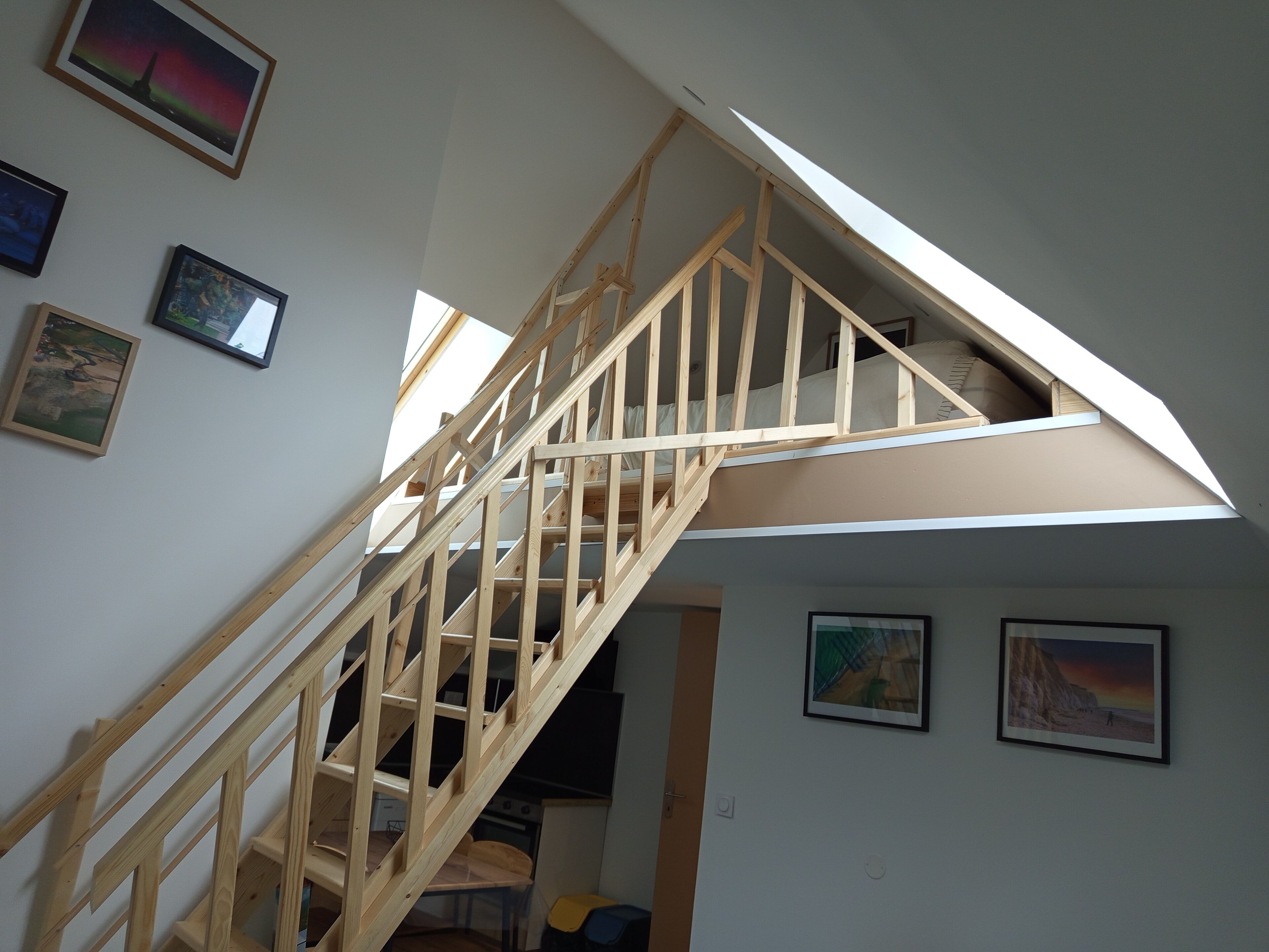 A wooden staircase leading to a loft bedroom with skylights, framed artwork on the walls, and a visible kitchen area below.