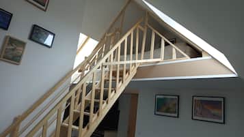 A wooden staircase leading to a loft bedroom with skylights, framed artwork on the walls, and a visible kitchen area below.