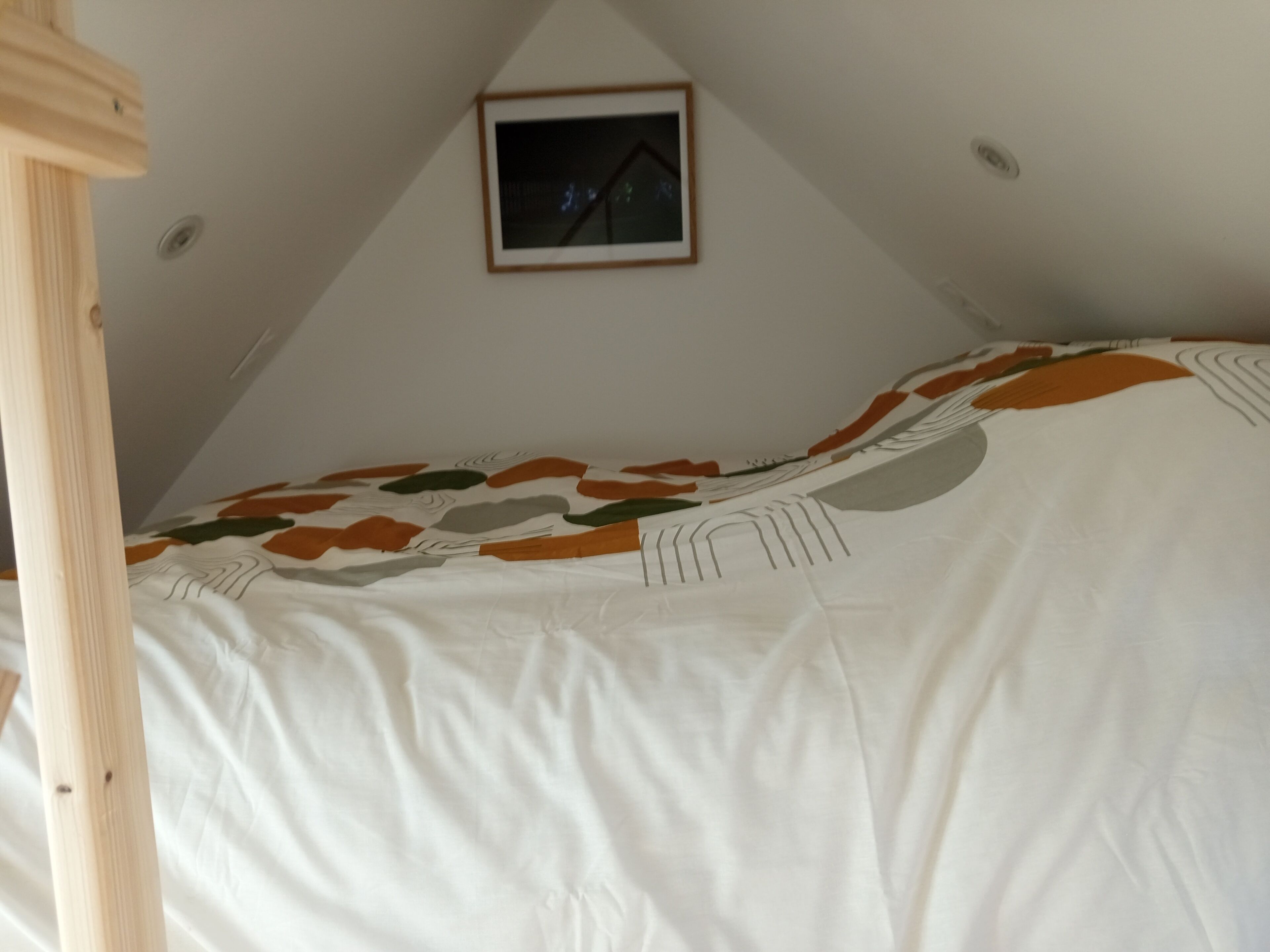A bedroom with a bed covered in a white blanket with leaf patterns, a wooden headboard, and a framed picture on the wall.