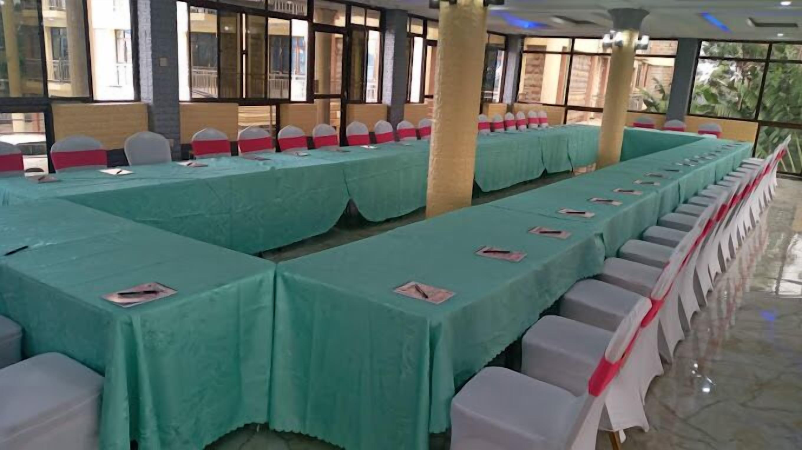 A conference room with long tables covered in green tablecloths, white chairs with red sashes, and a glass partition wall.