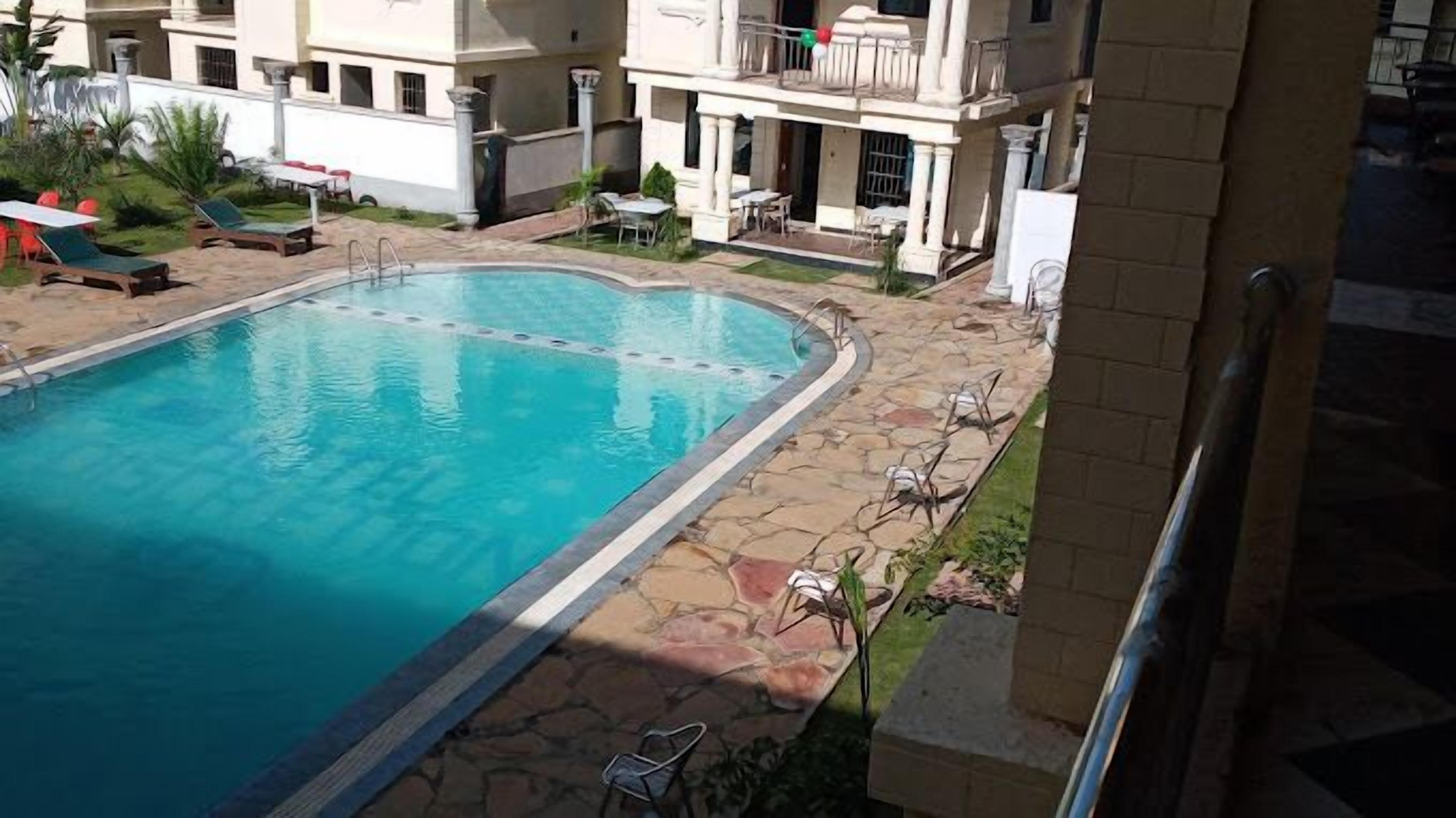 A swimming pool with a stone deck, lounge chairs, and a building with balconies in the background.