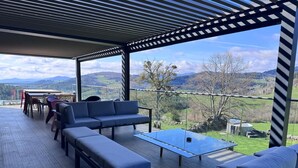 A covered outdoor seating area with a sofa, benches, and a table, overlooking a scenic landscape.