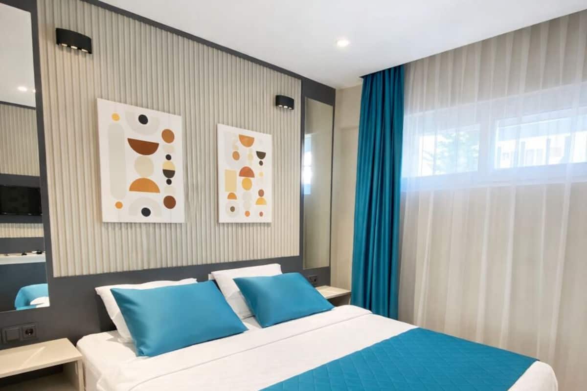 A modern hotel room with a large bed, a bedside table, a mirror, and abstract wall art.