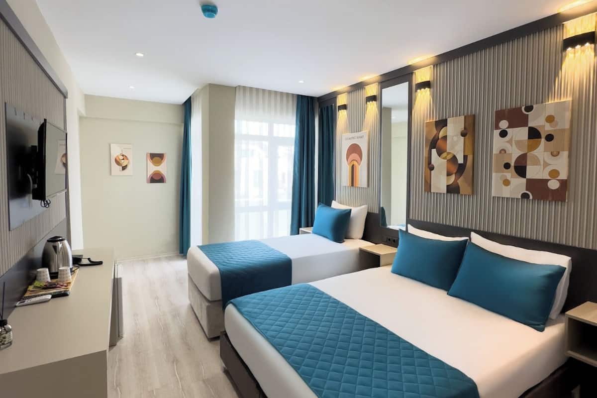 A modern hotel room with two beds, a flat-screen TV, a desk with a kettle, and artwork on the walls.