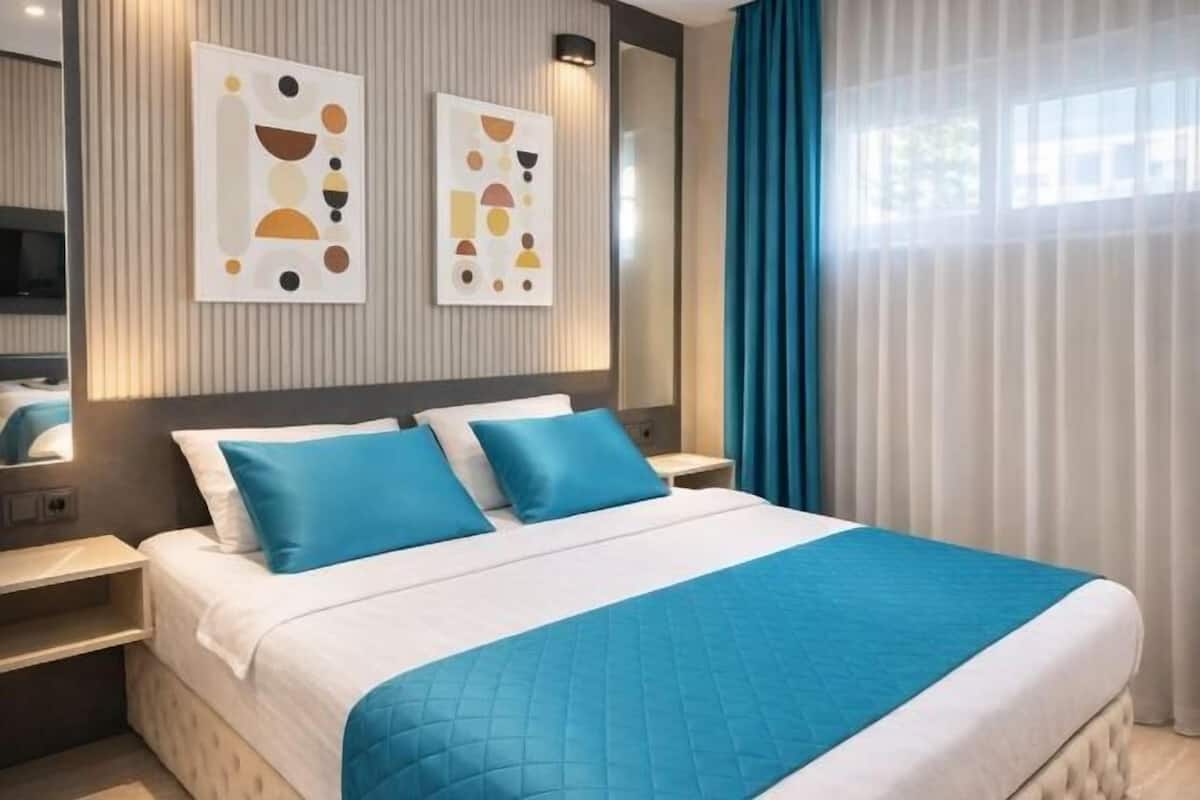 A modern hotel room with a large bed, two blue pillows, a white quilted bedspread, and abstract wall art.