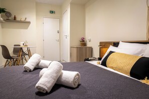 A neatly made bed with rolled towels, a bedside table with a lamp, a small plant, and a wooden headboard. - Nobia 5 (Athens)