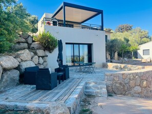 A modern two-story building with a stone facade, outdoor seating area, and a view of the surrounding landscape.