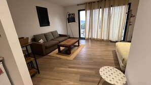 A modern living room with a sofa, coffee table, and a small round table. There is a large window with curtains and a view of the outdoors. - Large 50m2 studio, in the heart of the Maquis, San gavino di Figari (San gavinu Di figari)