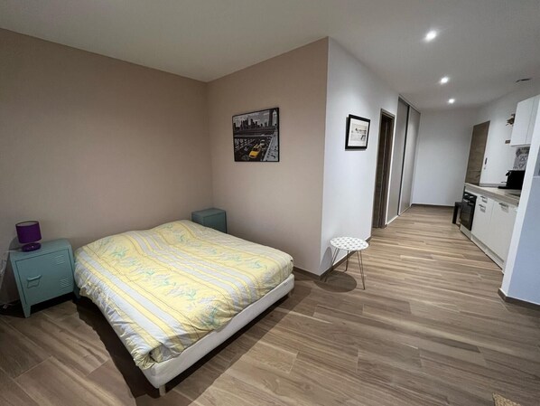 A bedroom with a bed, a small bedside table, a nightstand, a wall-mounted picture, and a hallway leading to a kitchenette.