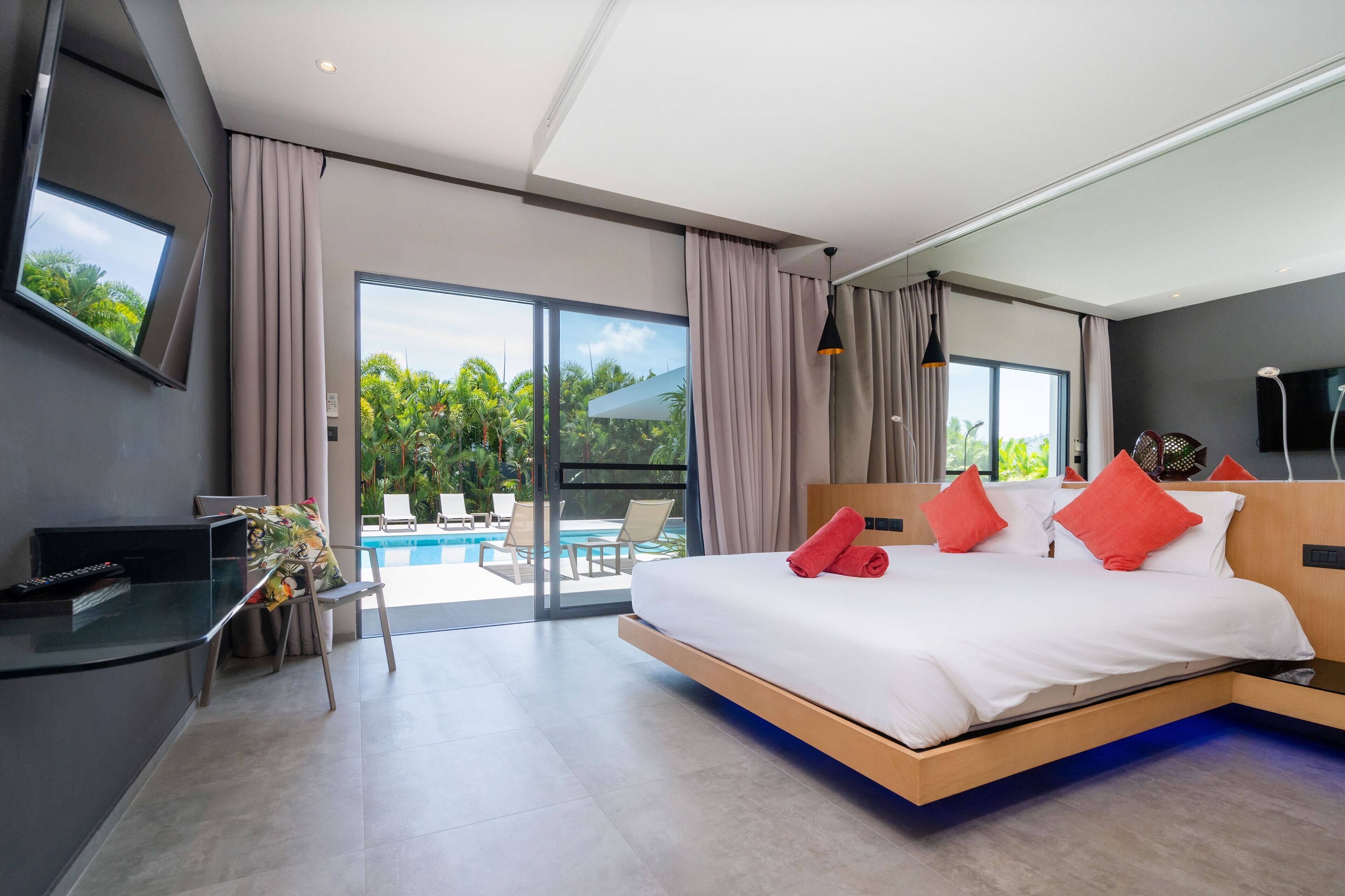 A modern hotel room with a large bed, a desk, a TV, and a view of the pool and greenery outside.