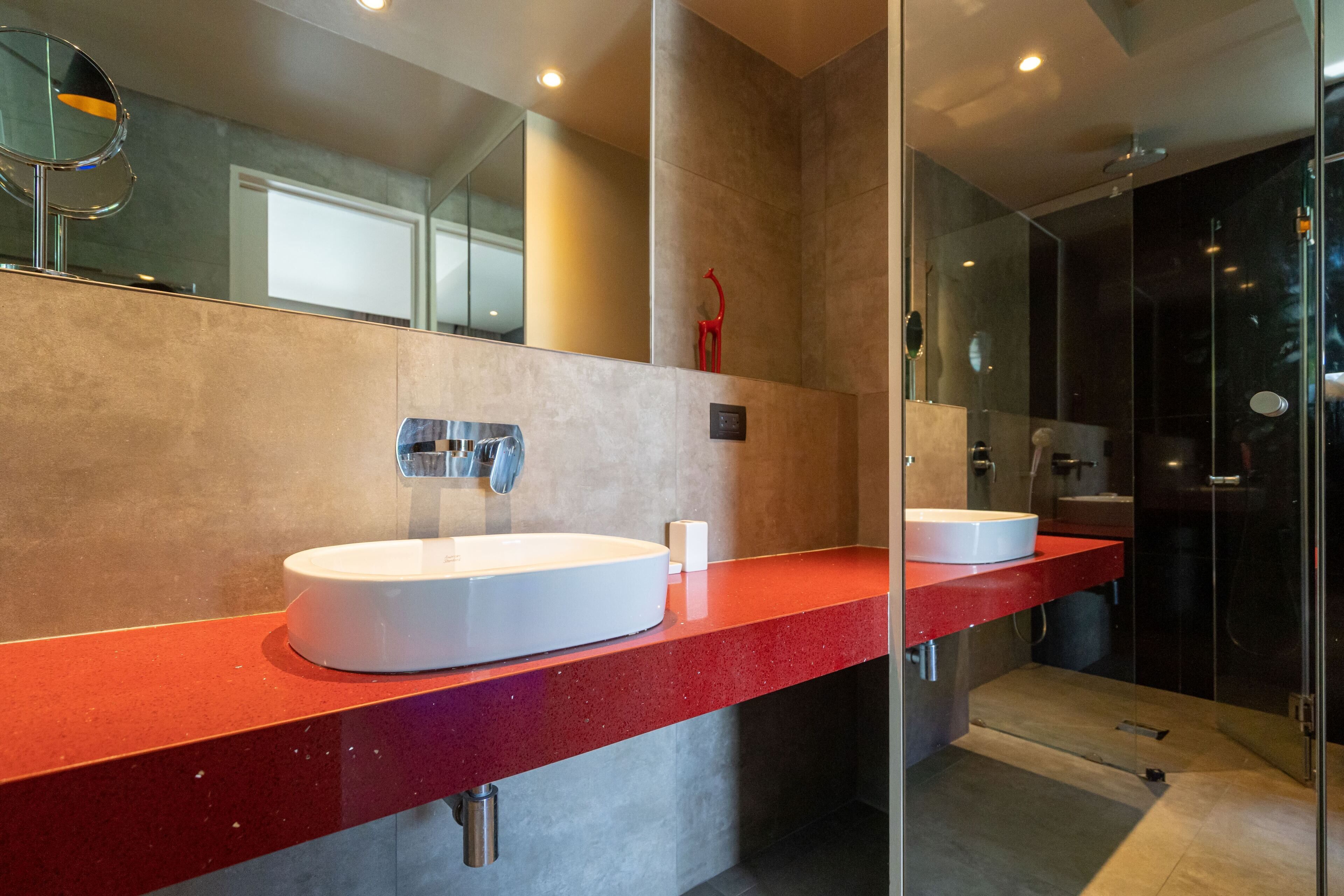 Un baño moderno con encimera roja, dos lavamanos blancos, un espejo grande y una cabina de ducha con paredes de vidrio.